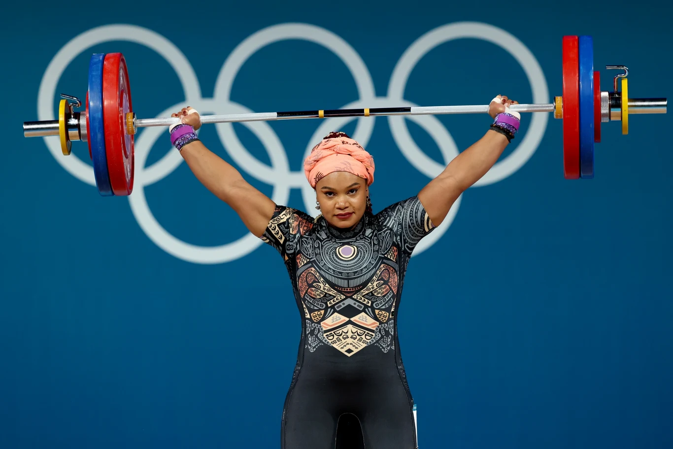 kobieta w sportowym stroju i opasce na głowie unosi nad głową sztangę z dużymi ciężarami, w tle widoczne olimpijskie koła