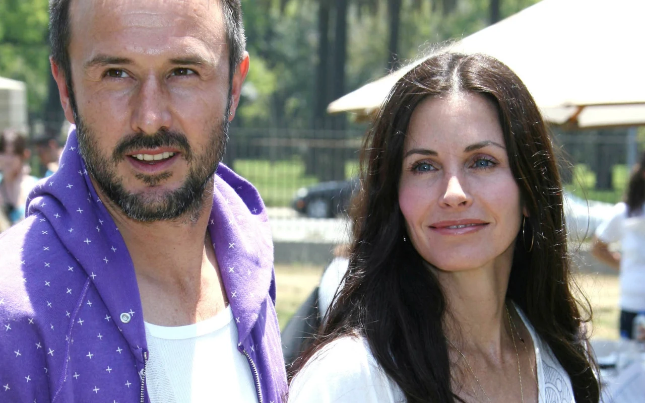 Dwoje ludzi stoi obok siebie na zewnątrz, mężczyzna w fioletowej rozpinanej bluzie z białym podkoszulkiem, kobieta z długimi, ciemnymi włosami ubrana na jasno, oboje patrzą w nieco innym kierunku, w tle elementy parku i rozmyty namiot.