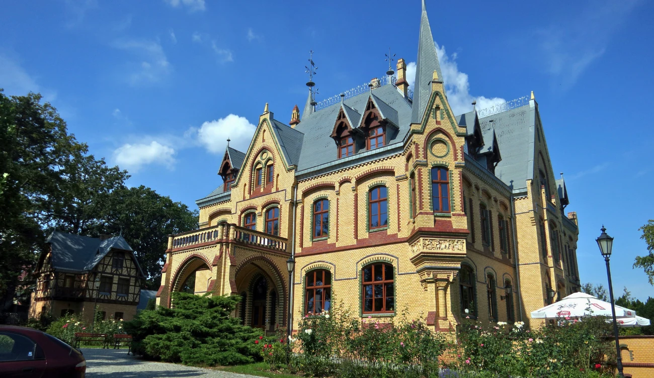 okazały, żółty pałac z czerwonymi detalami i stromym dachem, otoczony bujną zielenią, po lewej stronie zabytkowy budynek gospodarczy, z przodu róże i latarnia, w tle błękitne niebo z chmurami