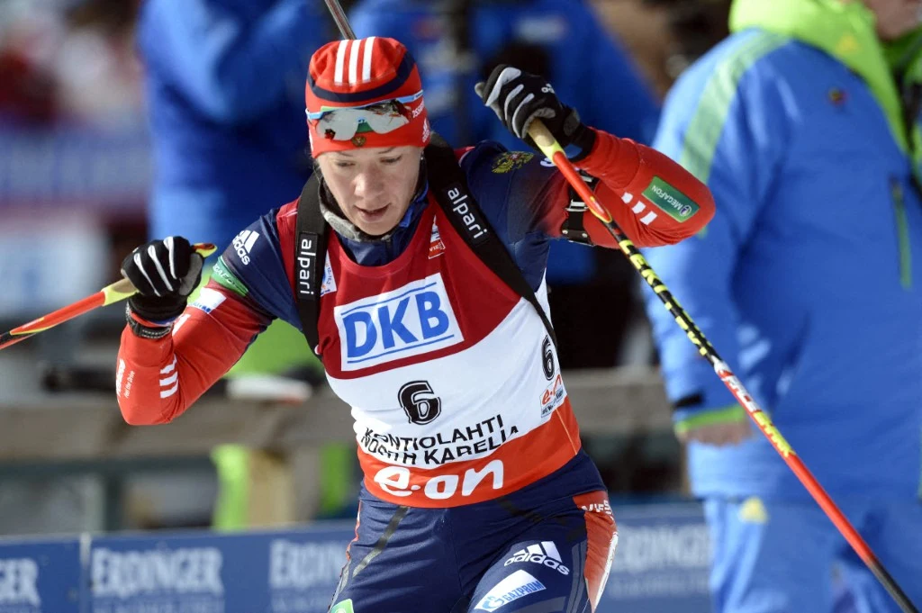 biathlonista w sportowym stroju i goglach na głowie, trzymający kijek narciarski, intensywnie skupiony podczas wyścigu, z widocznym numerem startowym na piersi, w tle inne osoby w sportowych ubraniach