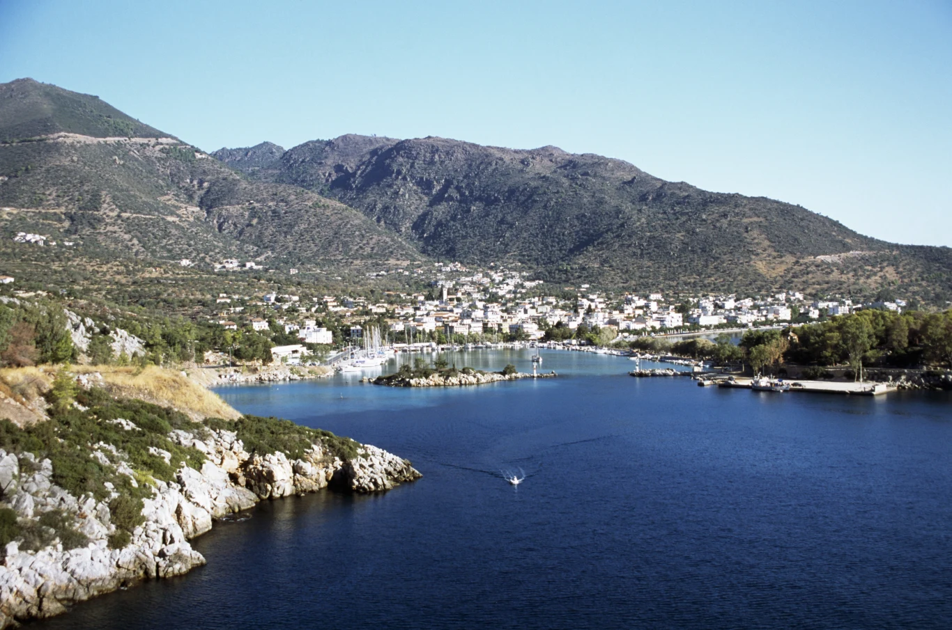 Nadmorska miejscowość położona u podnóża gór, z licznymi białymi budynkami otaczającymi zatokę; na wodzie widoczna płynąca łódka oraz mała marina z zacumowanymi jachtami.