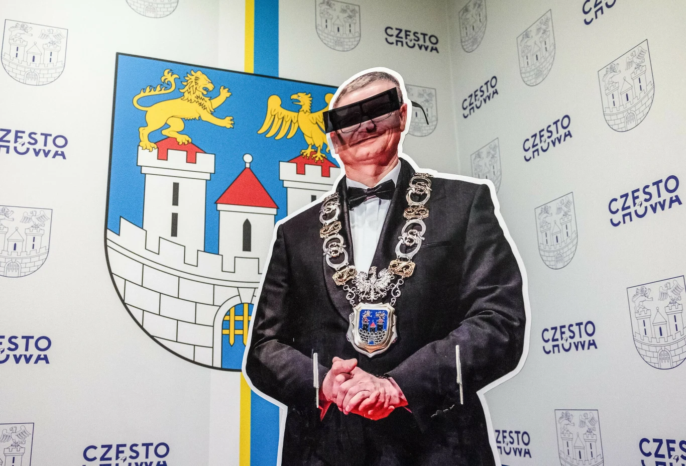 Zarzuty dla prezydenta Krzysztofa Matyjaszczyka. Referendum w Częstochowie Tekturowa postać w garniturze z łańcuchem prezydenckim, oczy zasłonięte paskiem, na tle herbu Częstochowy i logotypów miasta.