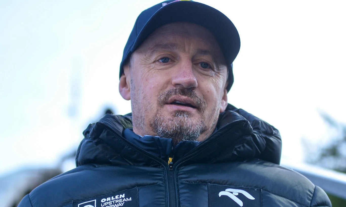 Adam Małysz Mężczyzna w granatowej kurtce sportowej z logotypami oraz granatowej czapce z daszkiem, patrzący przed siebie na tle jasnego nieba.