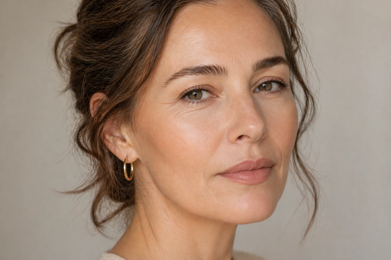 kobieta o jasnej cerze i brązowych włosach spiętych w luźny kok, nosząca złote kolczyki koła, z delikatnym makijażem i spokojnym, pewnym siebie wyrazem twarzy