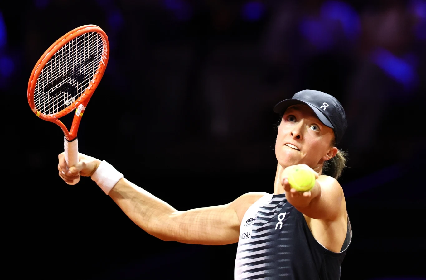 Tenisistka w ciemnej czapce i stroju sportowym przygotowuje się do serwisu, skupiając wzrok na piłce unoszonej do góry, z rakietą uniesioną wysoko nad głową.