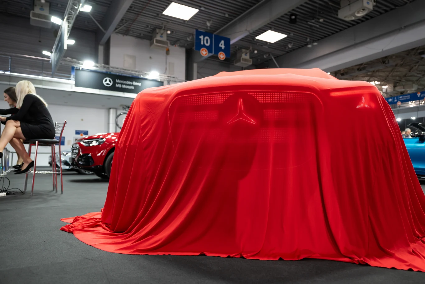 Honoru europejskich producentów broni Mercedes samochód przykryty czerwonym materiałem z wyraźnym zarysem grilla i logo Mercedesa, w tle fragmenty innych aut oraz postać kobiety siedzącej przy stole po lewej stronie
