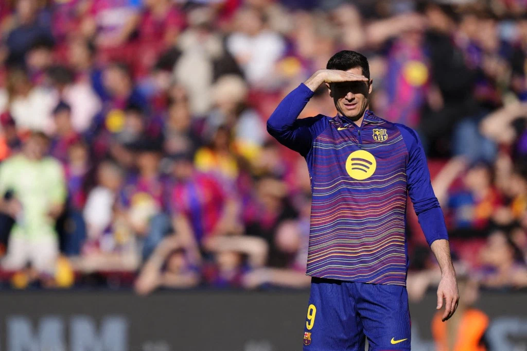 Piłkarz FC Barcelony w stroju treningowym z numerem 9 zasłania ręką oczy przed słońcem, stojąc na boisku podczas meczu, w tle rozmyta grupa kibiców na trybunach.