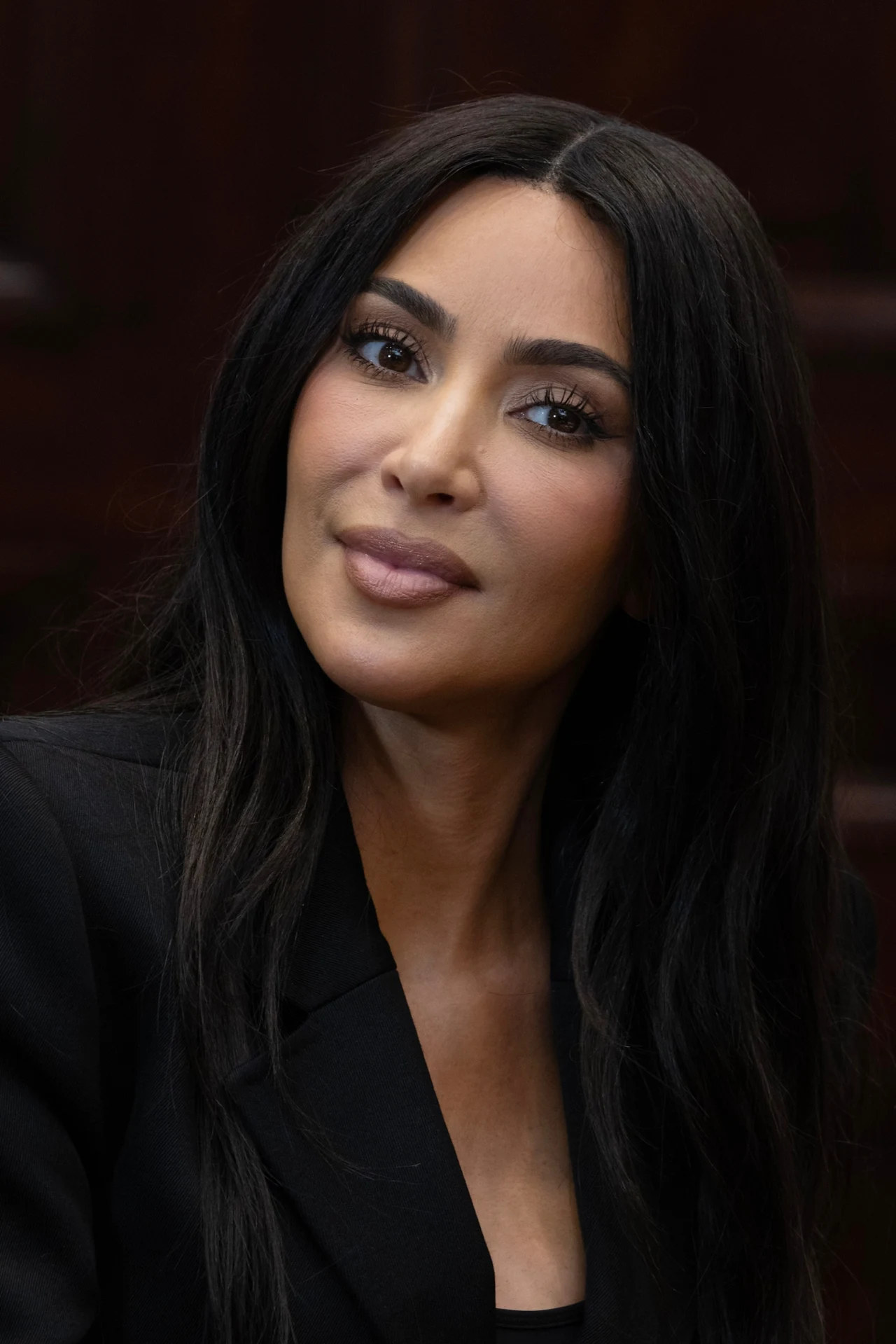 kobieta o długich, ciemnych włosach i wyrazistym makijażu, ubrana w czarną marynarkę, spoglądająca lekko na bok z delikatnym uśmiechem