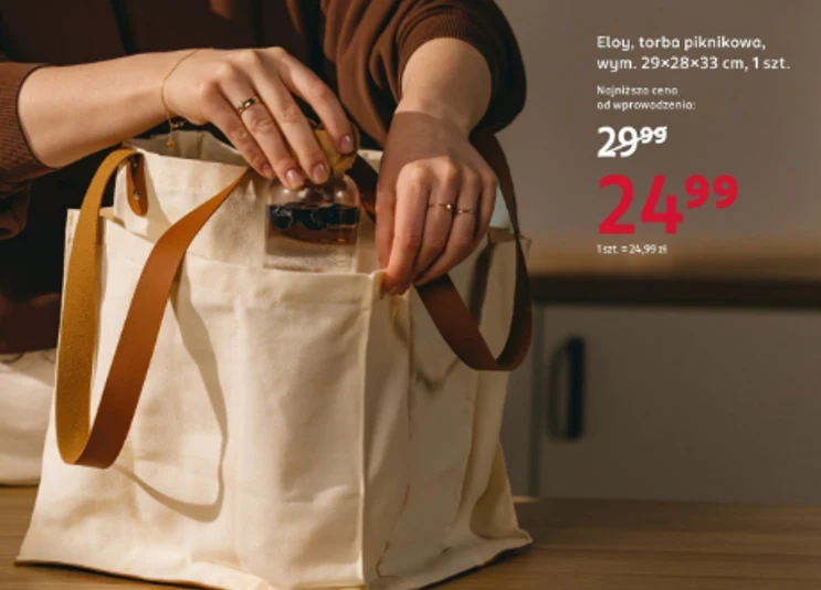 Torba piknikowa za 24,99 zł – sprawdzi się na spacer, piknik i zakupy Duża, kremowa torba piknikowa z brązowymi uchwytami stoi na drewnianym blacie, a osoba wkłada do środka przezroczystą saszetkę; na torbie widoczne są detale produktu i cena promocyjna.