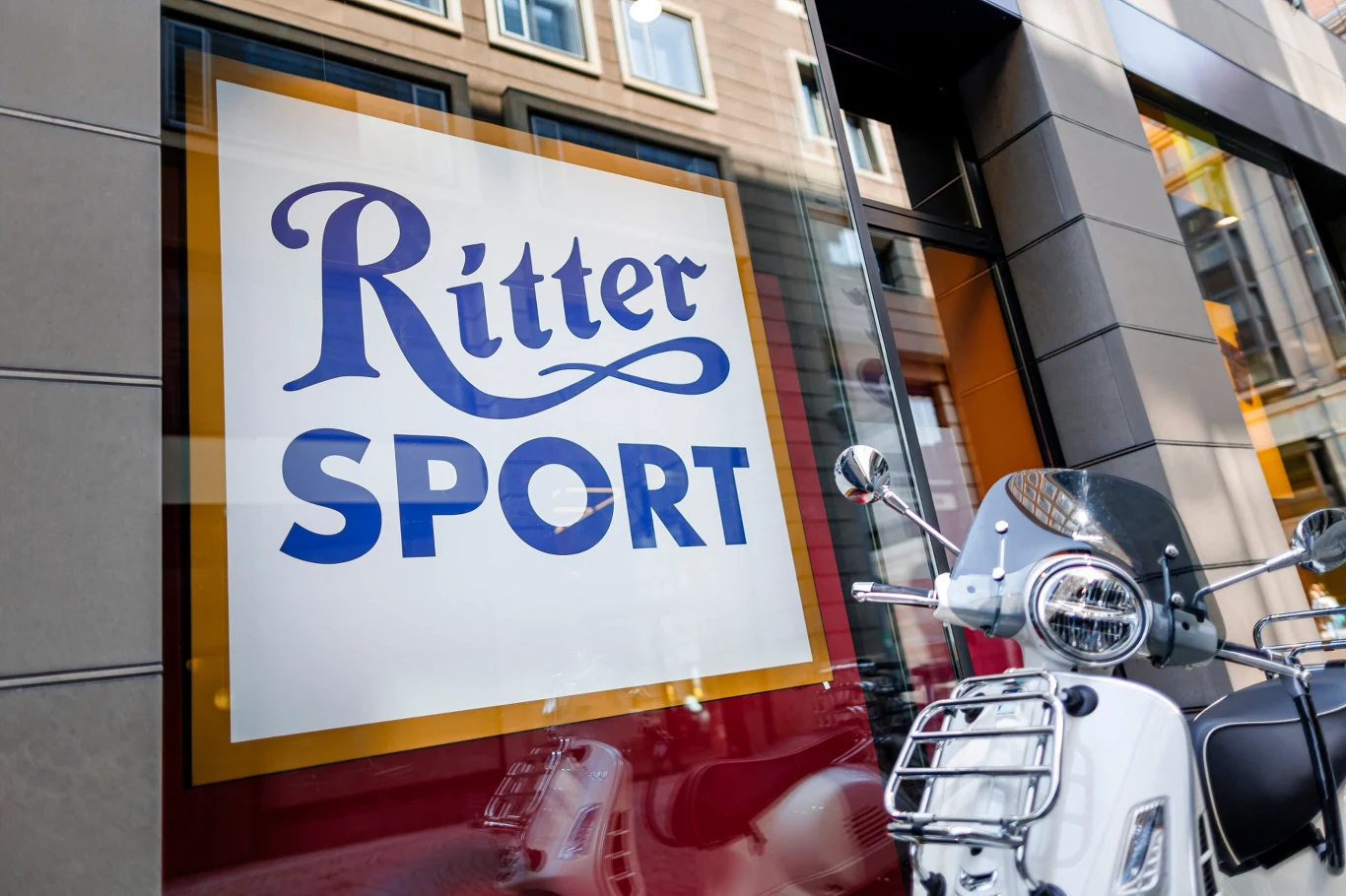 Ritter Sport tego jeszcze nie robił. Pierwsze zwolnienia od 110 lat Logo Ritter Sport na witrynie sklepu, obok zaparkowany skuter odbijający się w szybie