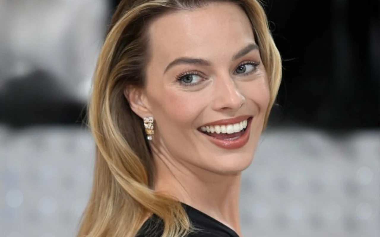 uśmiechnięta kobieta z długimi blond włosami, elegancko ubrana, patrzy przez ramię na tle rozmytego, jasnego tła