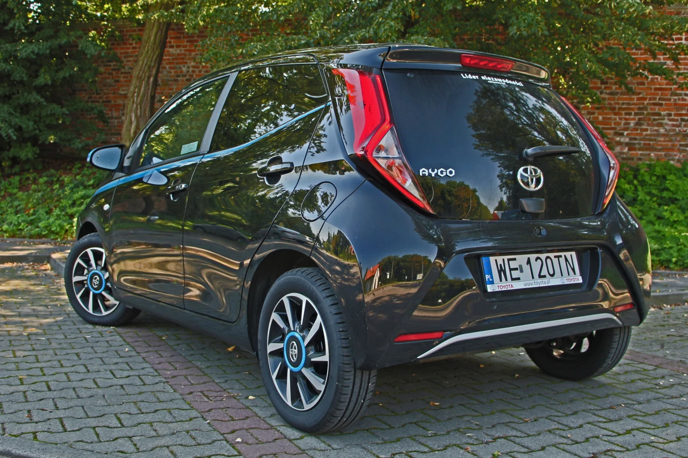 Technicznie to bliski krewny Citroena C1 i Peugeota 108, bo całe trio powstawało w czeskim Kolinie. czarny samochód osobowy Toyota Aygo z nowoczesnymi reflektorami tylnymi w kształcie litery X, stojący na parkingu brukowanym obok drzewa i ceglanego muru