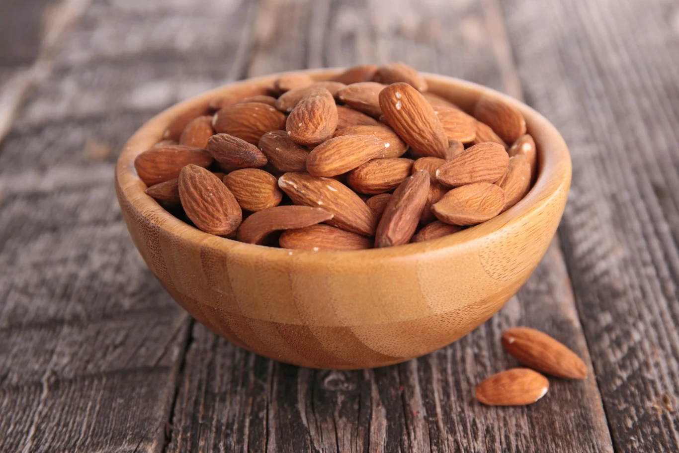 drewniana miska wypełniona całymi, niełuskanymi migdałami ustawiona na surowych, drewnianych deskach, kilka migdałów leży obok miski