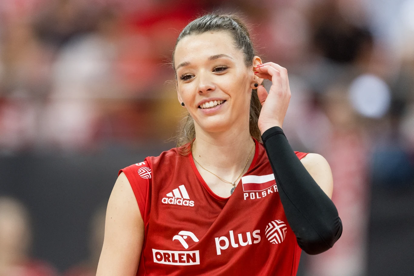 Zuzanna Górecka siatkarka w czerwonym stroju z logotypami sponsorów i napisem „Polska”, uśmiechnięta, z jednym rękawem sportowym, na hali sportowej
