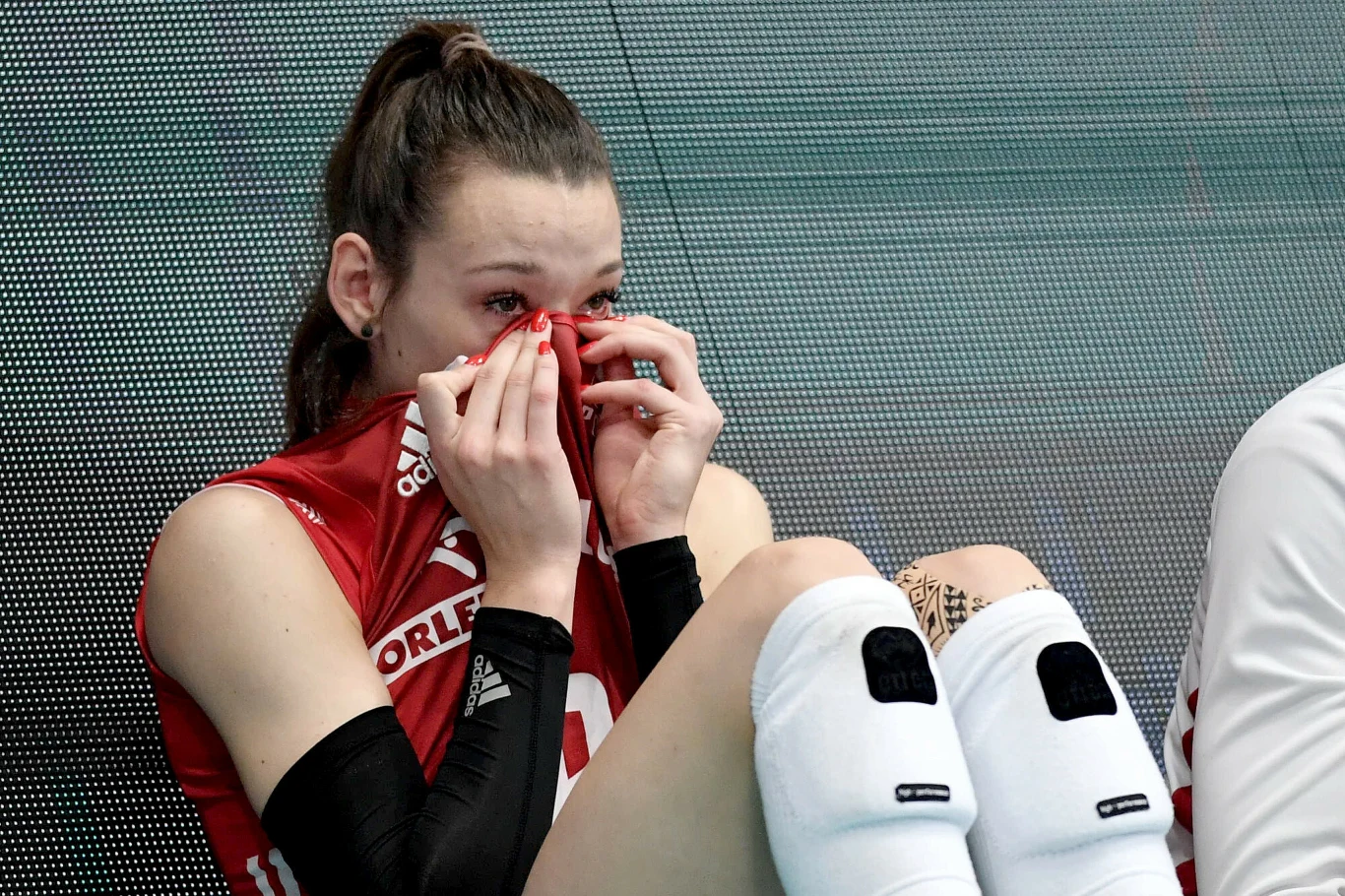 Zuzanna Górecka siatkarka w czerwonym stroju siedząca ze zgiętymi nogami, zasłaniająca twarz koszulką, wyrażająca smutek lub rozczarowanie