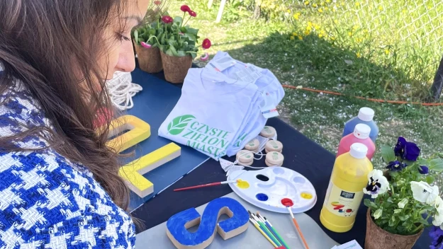 kobieta maluje drewniane litery farbami, na stole leżą kolorowe farby, pędzle, paleta malarska, sadzonki kwiatów w doniczkach oraz koszulka z napisem 'CASSIE SHOW MASA'