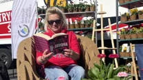 kobieta w okularach przeciwsłonecznych siedzi na kartonowym fotelu i czyta książkę, przed nią stół z owocami oraz doniczkami z kwiatami, w tle stoiska z roślinami i baner fundacji ekologicznej