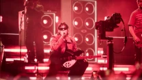 muzyk w okularach przeciwsłonecznych i czerwonej kurtce klęczy na scenie z gitarą elektryczną, wokół niego intensywne czerwone światła i duże głośniki, w tle widać kamerzystę i innych członków zespołu