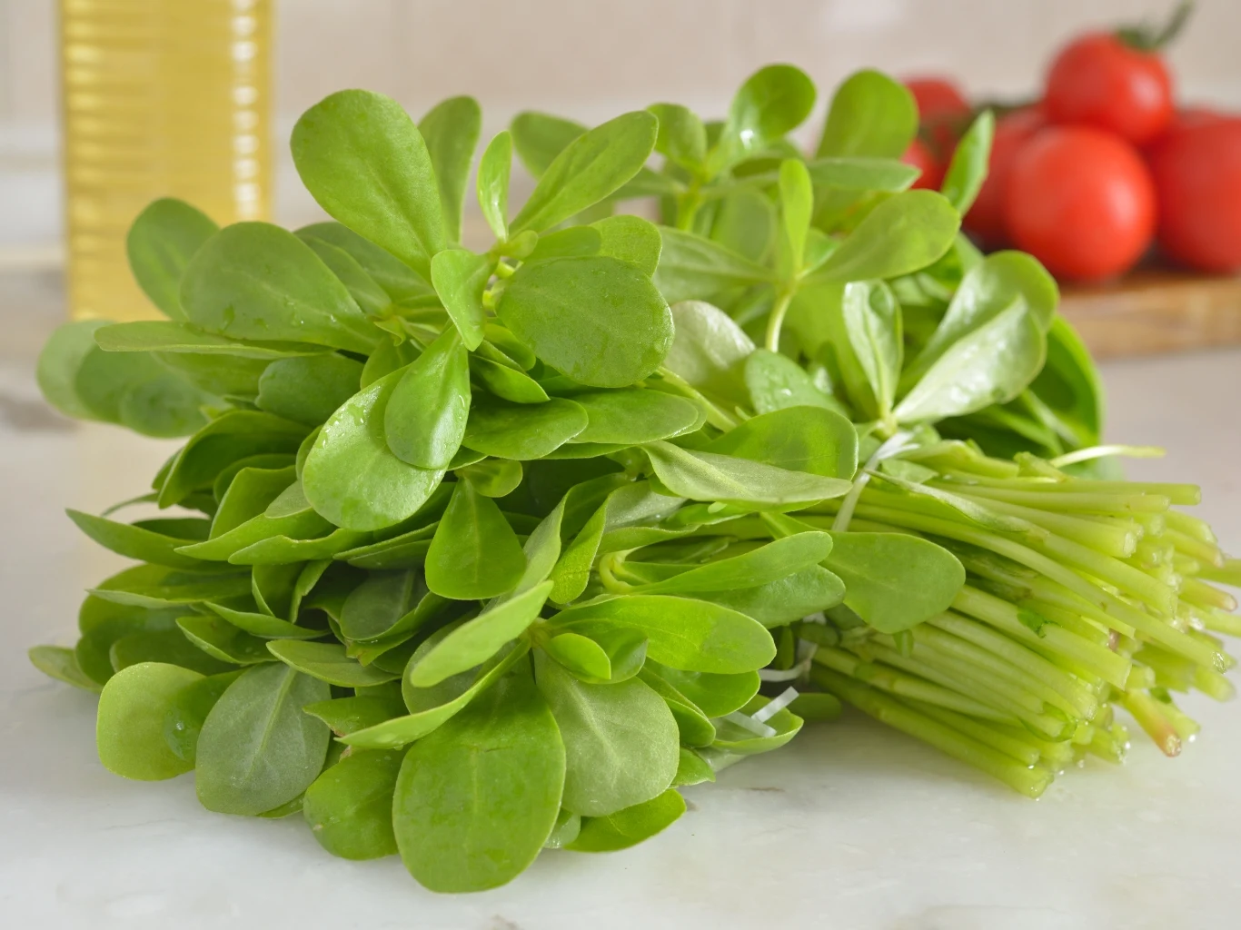 wiązka świeżego portulaku o jasnozielonych, błyszczących liściach ułożona na jasnym blacie kuchennym; w tle rozmyte czerwone pomidory oraz butelka oleju