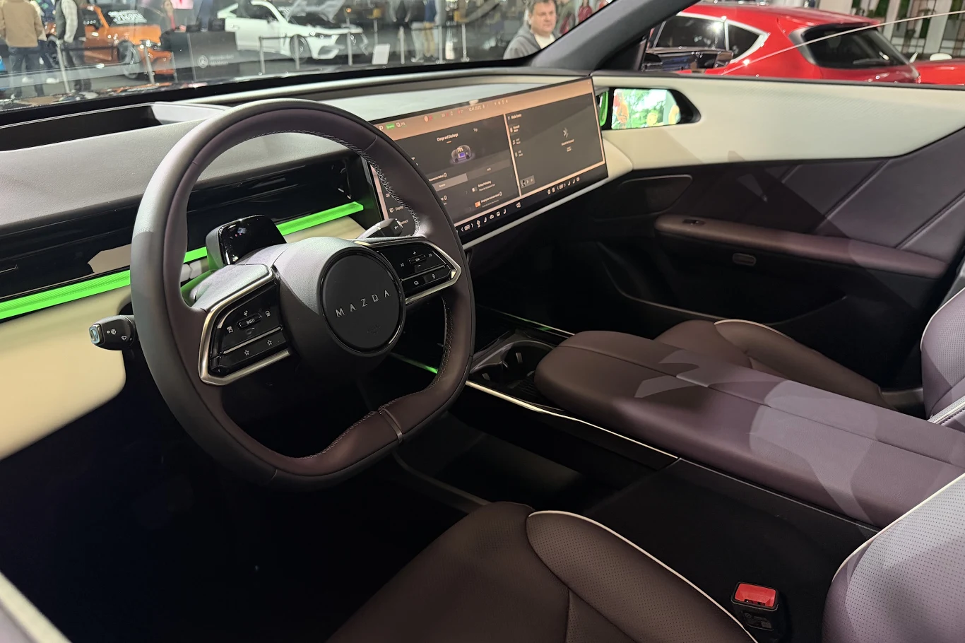 Mazda CX-6e nowoczesne wnętrze samochodu elektrycznego z minimalistyczną deską rozdzielczą, dużym panoramicznym ekranem dotykowym i podświetleniem LED w kolorze zielonym, kierownica o innowacyjnym kształcie oraz skórzane, wygodne fotele kierowcy i pasażera