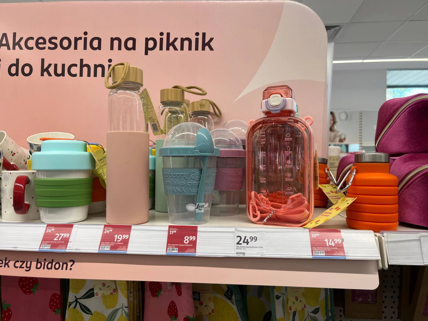 Pojemnik z łyżeczką – 8,99 zł Idealny na szybkie śniadanie w pracy lub na uczelni Kolorowe bidony, kubki i butelki wielokrotnego użytku ustawione na półce sklepowej, każdy z ceną i etykietą; w tle fragmenty różowych kosmetyczek oraz rozłożone składane silikonowe kubki.