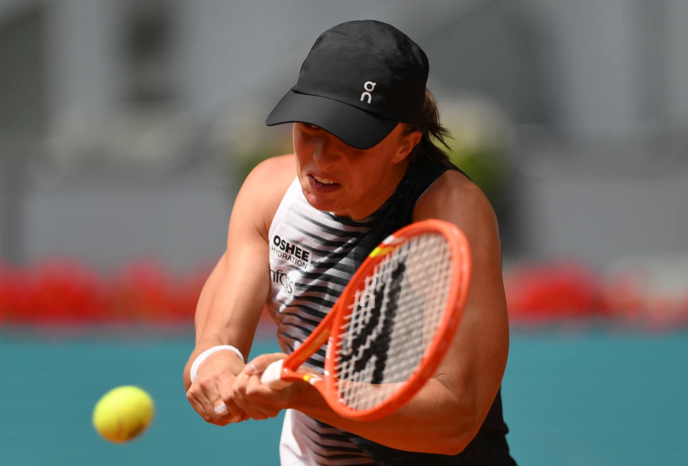 zawodniczka w czarnej czapce uderza żółtą piłkę tenisową dwuręcznym bekhendem, skupiona na odbiciu, w tle rozmyte trybuny