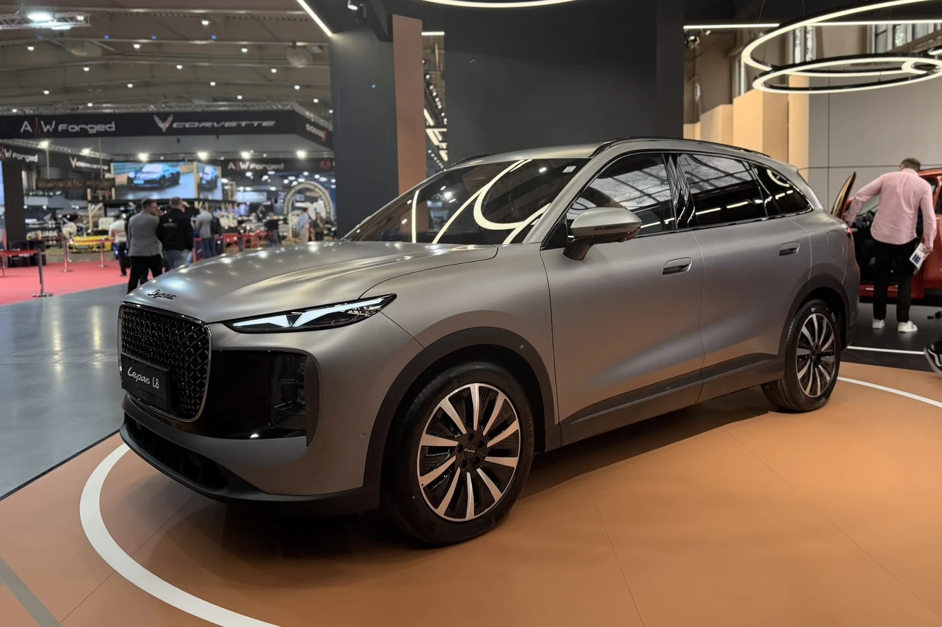 Lepas L8 nowoczesny SUV w matowym, szarym kolorze ustawiony na wyeksponowanym stanowisku targowym, z dużą atrapą chłodnicy, charakterystycznymi przednimi światłami LED oraz wysokimi, aluminiowymi felgami na tle efektownego, nowoczesnego oświetlenia wystawowego