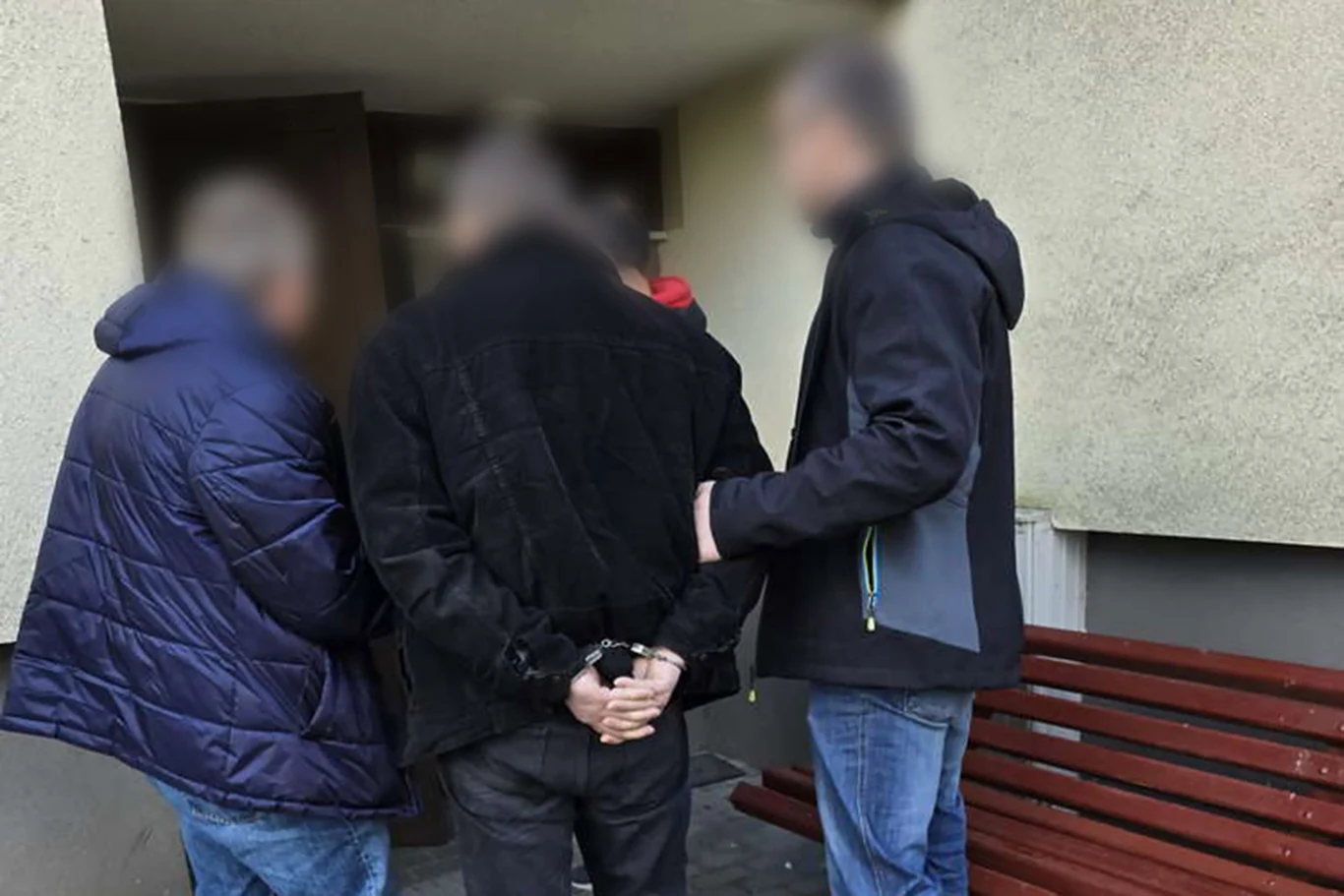 Wśród zatrzymanych znaleźli się dwaj diagności oraz handlarz samochodami | Fot. Policja Małopolska trzech mężczyzn stoi na zewnątrz budynku, jeden z nich ma skute kajdankami ręce za plecami, pozostali towarzyszą mu z obu stron