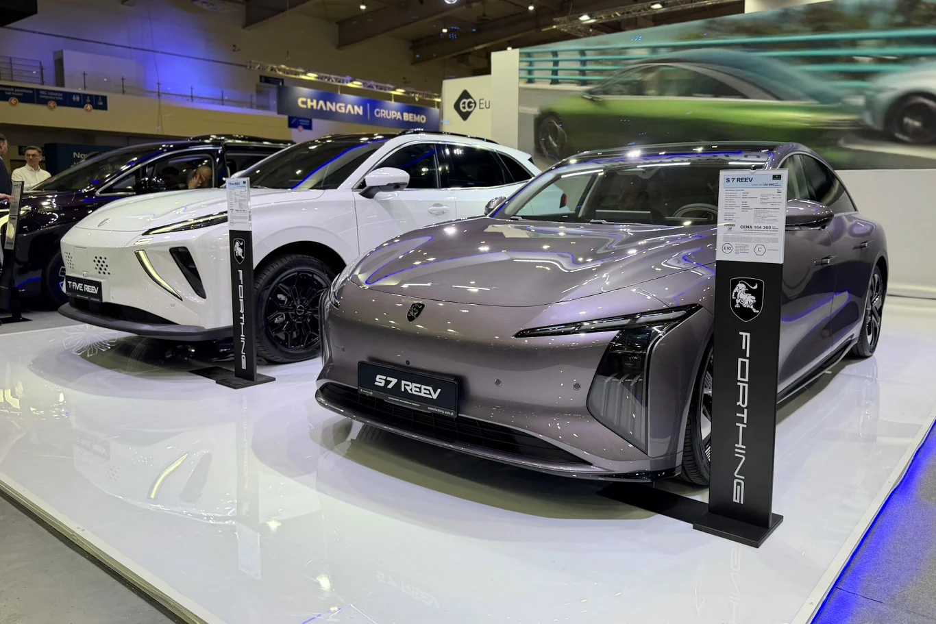 Forthing S7 i T-Five Forthing S7 REEV i T-Five na ekspozycji targowej; szary sedan S7 obok białego SUV-a, widoczne tabliczki informacyjne.
