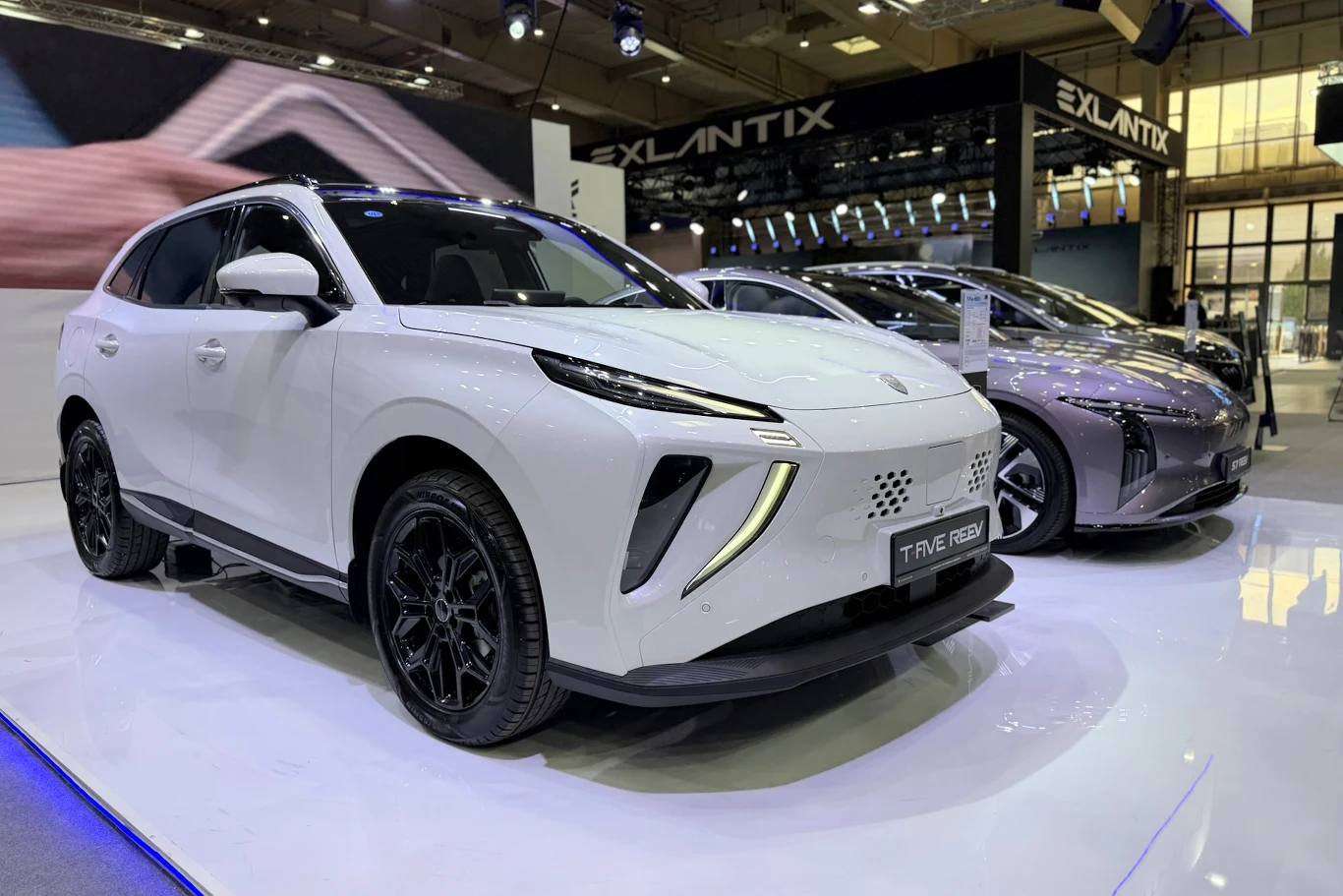 Forthing T-Five REEV dwa nowoczesne SUV-y elektryczne zaprezentowane na podświetlonym podium wystawowym, z dominującym białym modelem na pierwszym planie oraz futurystycznymi detalami świateł LED i aerodynamicznymi liniami nadwozia