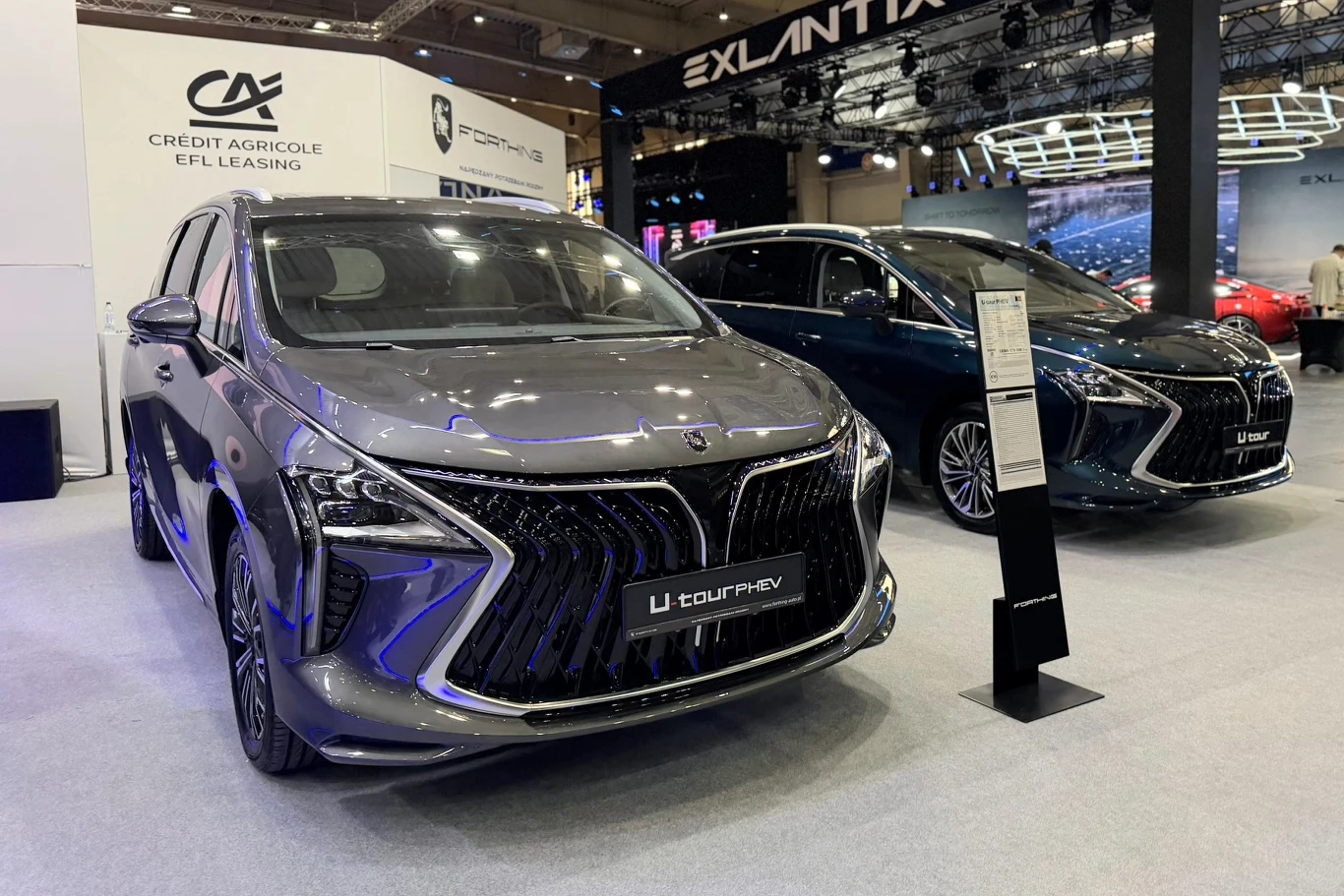 Forthing U-Tour PHEV dwa nowoczesne minivany o dynamicznej sylwetce z charakterystycznymi, szerokimi grillami i smukłymi reflektorami, ustawione na ekspozycji targowej z futurystycznym oświetleniem, informacyjną tablicą oraz logo sponsora w tle