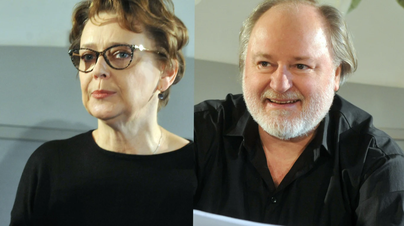 Dwie osoby w średnim wieku ubrane w ciemne ubrania, kobieta w okularach patrząca na wprost, mężczyzna z brodą i uśmiechem trzyma kartkę papieru.