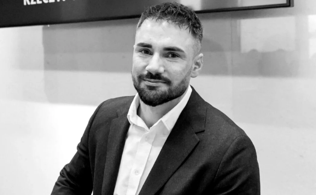 Mężczyzna z krótkimi ciemnymi włosami i brodą, ubrany w elegancką marynarkę oraz białą koszulę, siedzi przodem do obiektywu na tle jasnej ściany.