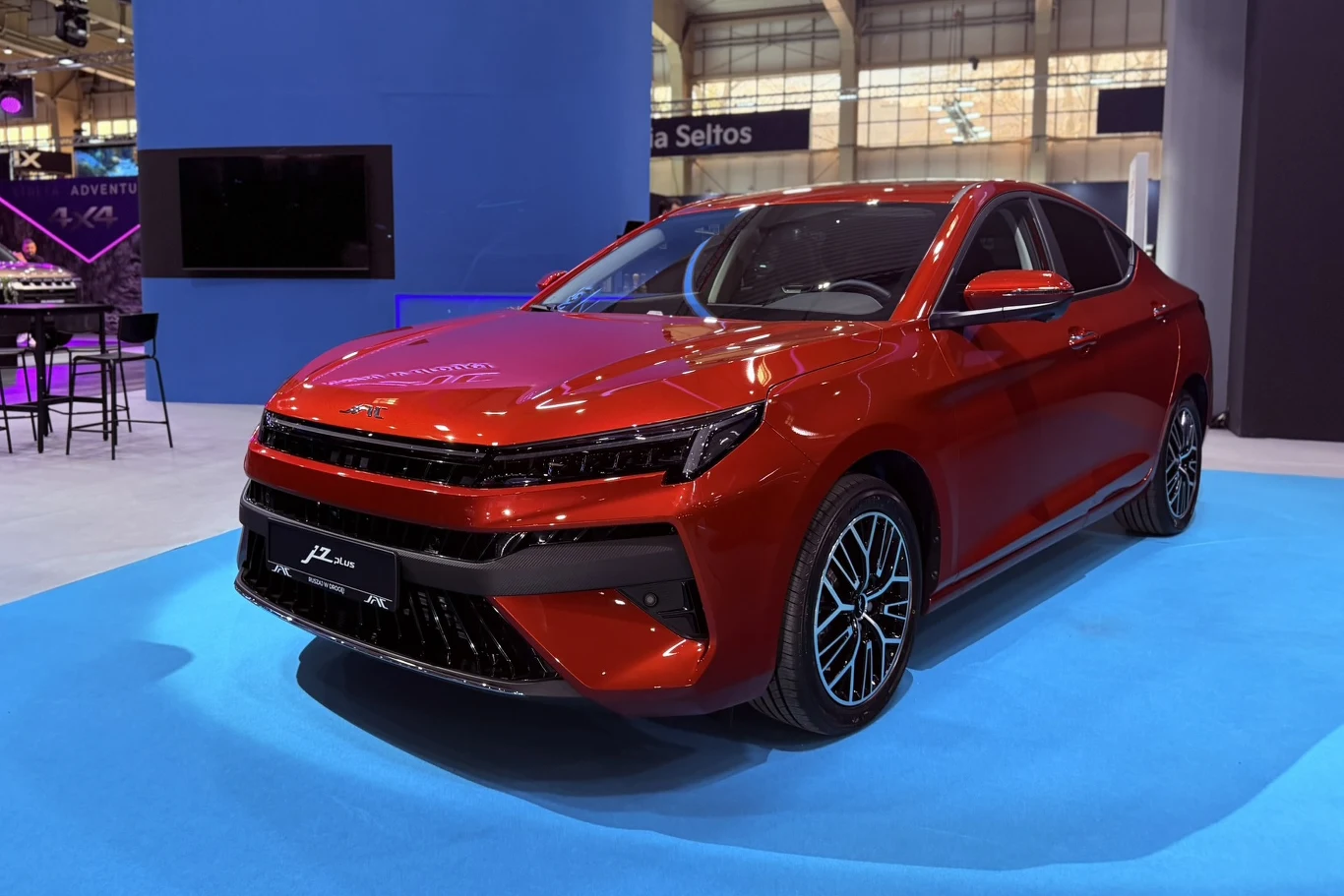 JAC J7 Plus czerwony, sportowy sedan z nowoczesnym designem, wyrazistym przednim grillem i czarnymi detalami, ustawiony na jasnoniebieskim podwyższeniu podczas targów motoryzacyjnych