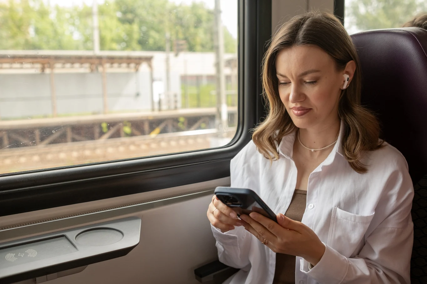 Kobieta z długimi, jasnobrązowymi włosami siedzi przy oknie w pociągu, trzyma smartfon w dłoniach i korzysta ze słuchawek bezprzewodowych, spoglądając z lekkim uśmiechem na ekran telefonu.