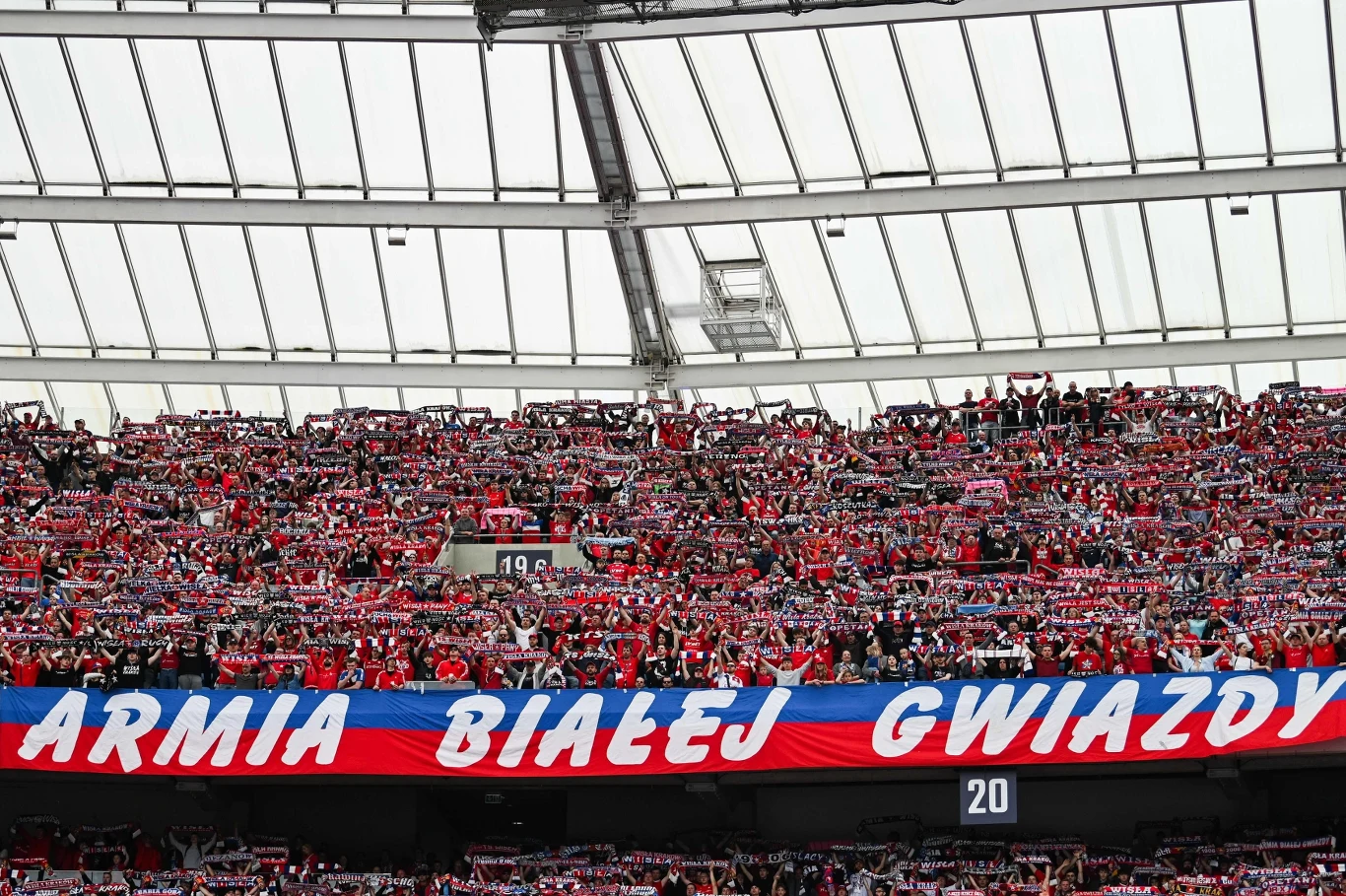 tłum kibiców ubranych w czerwone koszulki i szaliki, trzymających nad głowami barwy klubowe oraz dużą flagę z napisem 'ARMIA BIAŁEJ GWIAZDY' w stadionowej trybunie