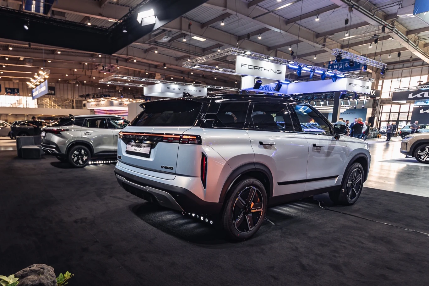 Jaecoo 7 Hybrid luksusowy SUV o srebrnym nadwoziu z kontrastującym czarnym dachem prezentowany na targach motoryzacyjnych, otoczony innymi nowoczesnymi samochodami i oświetleniem wystawowym