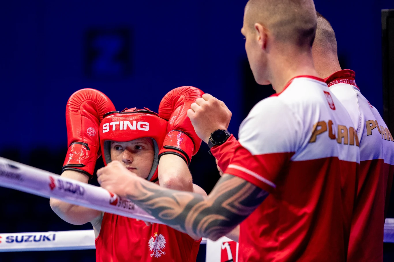 Bokser w czerwonym stroju z orzełkiem na piersi stoi w ringu, mając uniesione ręce w rękawicach bokserskich oraz kasku ochronnym, podczas gdy trener w biało-czerwonej koszulce z dużym napisem 'POLSKA' udziela instrukcji, prezentując gesty dłonią.