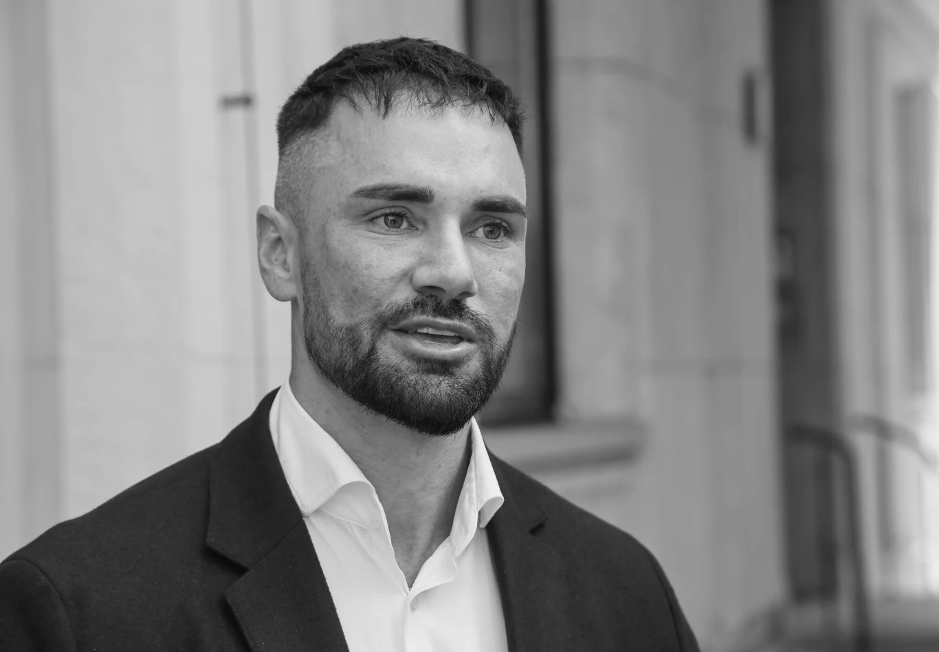 Mężczyzna z zarostem w garniturze i białej koszuli stoi przed jasnym, rozmytym tłem przypominającym elewację budynku.