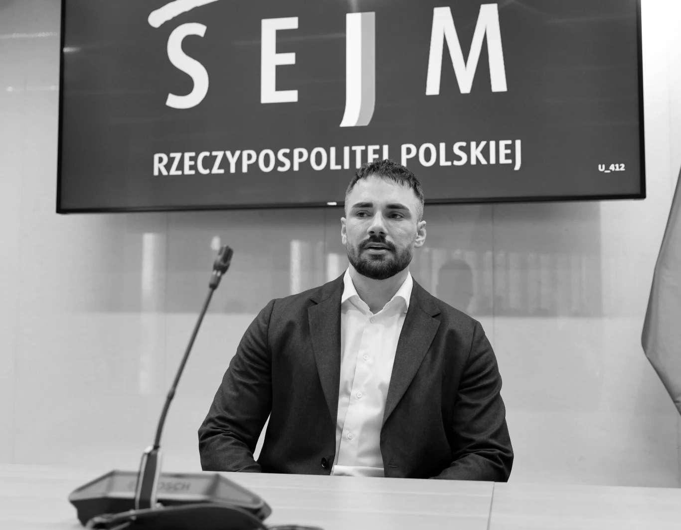 mężczyzna w marynarce i białej koszuli siedzi za stołem konferencyjnym, przed mikrofonem, na tle ekranu z napisem 'SEJM RZECZYPOSPOLITEJ POLSKIEJ'