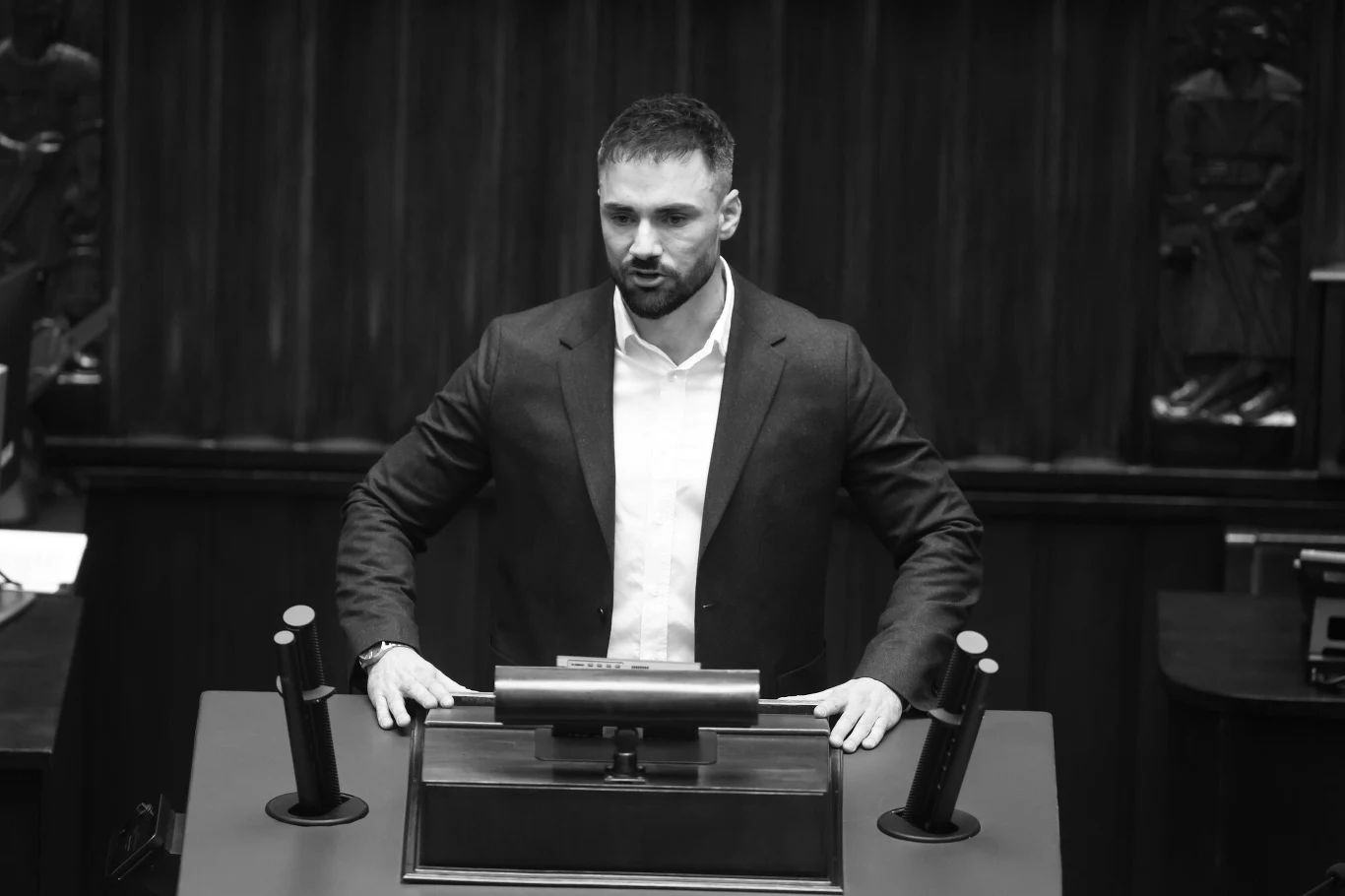 Łukasz Litewka nie żyje. Kondolencje rodzinie złożyły m.in. sosnowieckie kluby piłkarskie Mężczyzna w garniturze stoi za mównicą w sali przypominającej parlament lub inną oficjalną instytucję, ręce opiera na pulpicie, wyraz twarzy poważny.