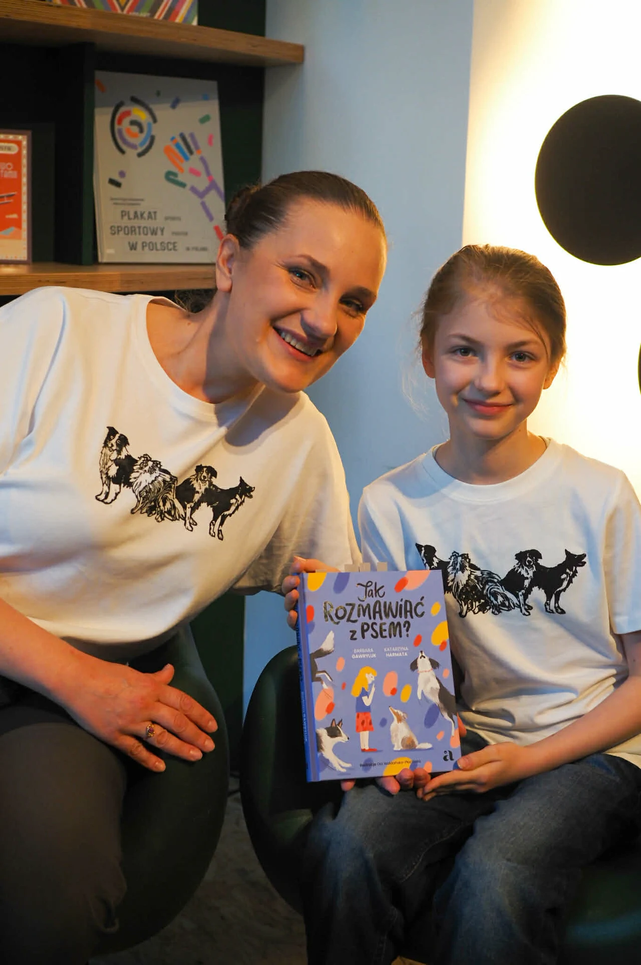 Zosia, córka Katarzyny Harmaty dziś ma 11 lat i jest niezwykle empatyczna w stosunku do zwierząt, a także wykazuje się niezwykłą wiedzą i dojrzałością w kontakcie z nimi Dwie uśmiechnięte osoby w jasnych koszulkach z czarnymi psami, siedzą obok siebie i trzymają książkę o psach pt. „Jak rozmawiać z psem?” w kolorowej okładce; w tle półka z książkami i plakatami.