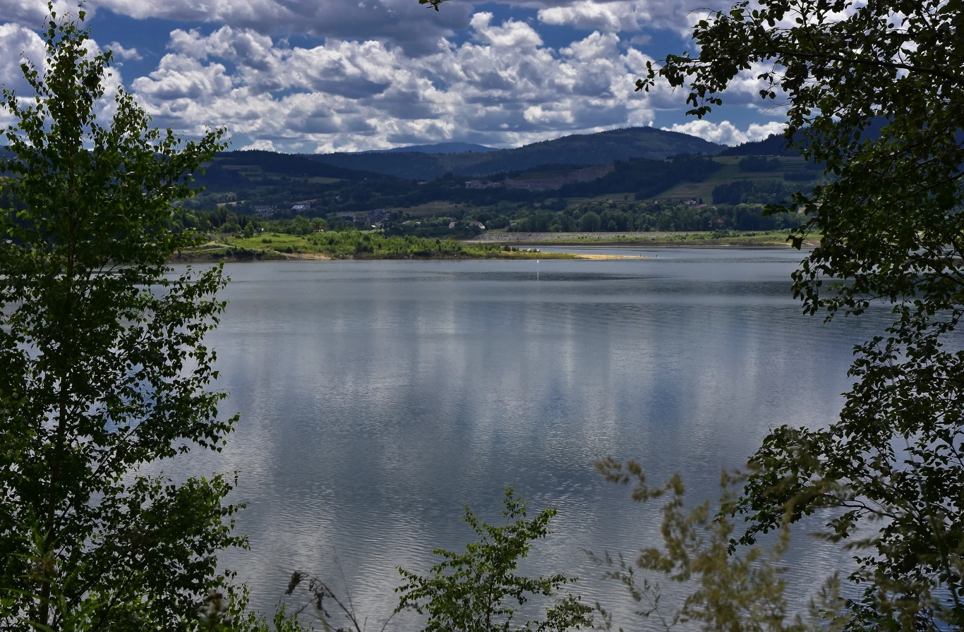 spokojna tafla jeziora otoczona zielenią drzew, w tle rozciągają się wzgórza porośnięte lasami pod pochmurnym, dynamicznym niebem
