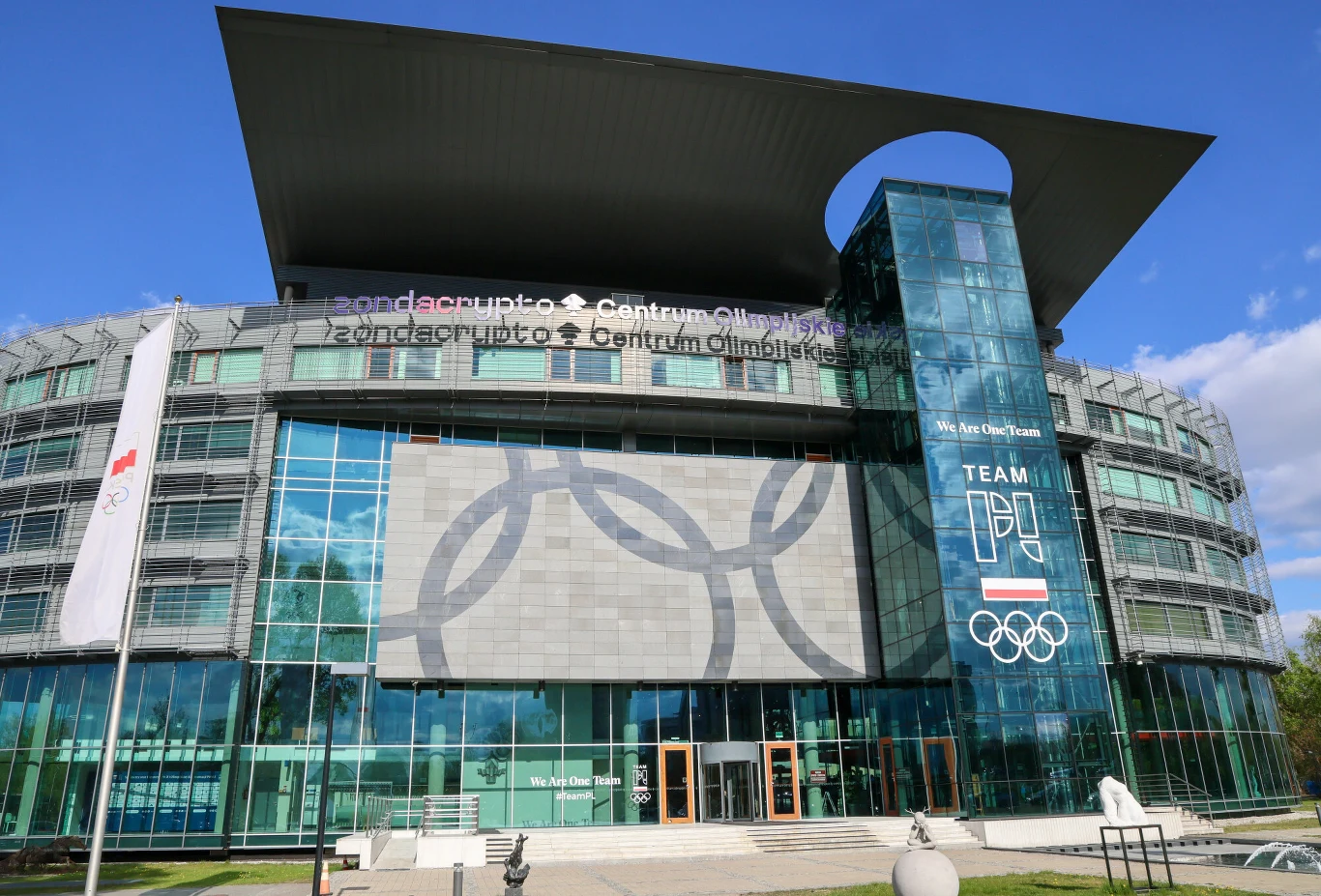nowoczesny budynek o przeszklonej fasadzie ze szklanym wejściem oraz dużym symbolem olimpijskich kół na froncie, obok widoczne logo Team Poland i polska flaga