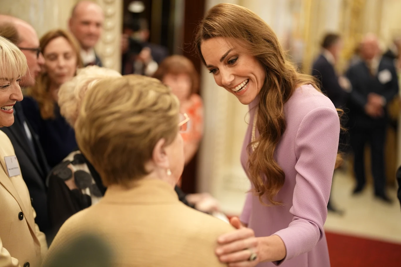 Księżna Kate uwielbia rozmawiać z poddanymi, ale są sytuacje, gdy jest to dla niej wymagające uśmiechnięta kobieta w liliowej sukience pochyla się i rozmawia z grupą starszych osób w eleganckim wnętrzu, kładąc rękę na ramieniu jednej z nich