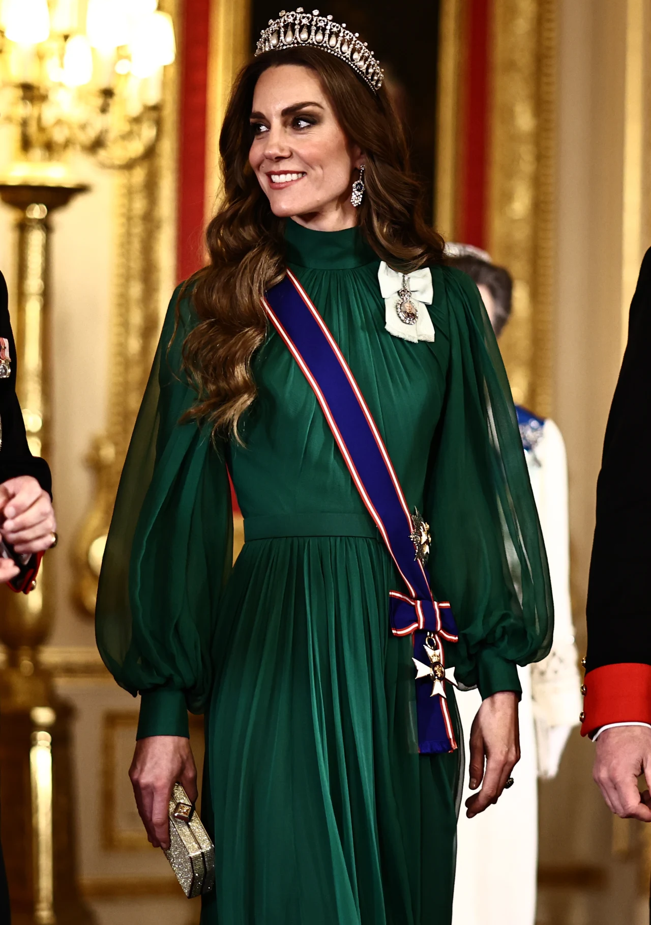 Księżna Walii od początku pełni swoją rolę w sposób godny podziwu kobieta w eleganckiej, zielonej sukni z bufkami, ozdobiona tiarą, orderem przewieszonym przez ramię oraz broszką, trzymająca w ręku złotą kopertówkę; otoczenie sugeruje pałacowe wnętrze z bogatymi dekoracjami