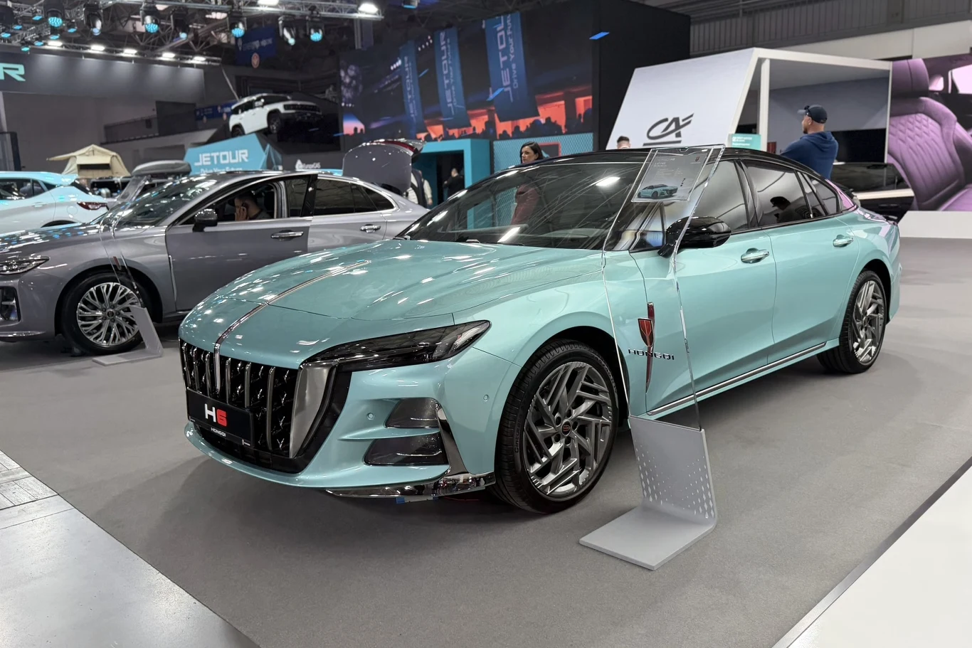 Hongqi H6 jasnoniebieski nowoczesny sedan z dużym, chromowanym grillem z przodu, wyrazistymi reflektorami oraz aerodynamicznymi felgami, ustawiony na ekspozycji targowej z otaczającymi go innymi samochodami i wystawową aranżacją w tle