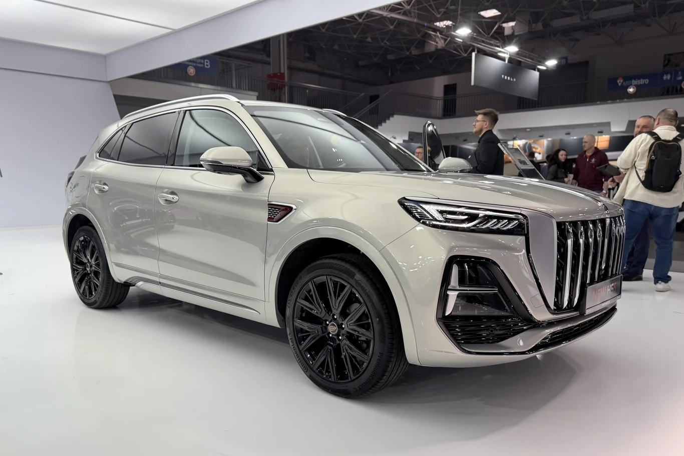 Hongqi HS5 biały, nowoczesny SUV o wyrazistym, masywnym grillu i futurystycznych, wąskich światłach, stojący na czarnych felgach w jasnym, przestronnym wnętrzu budynku wystawowego, wokół kilka osób przyglądających się pojazdowi