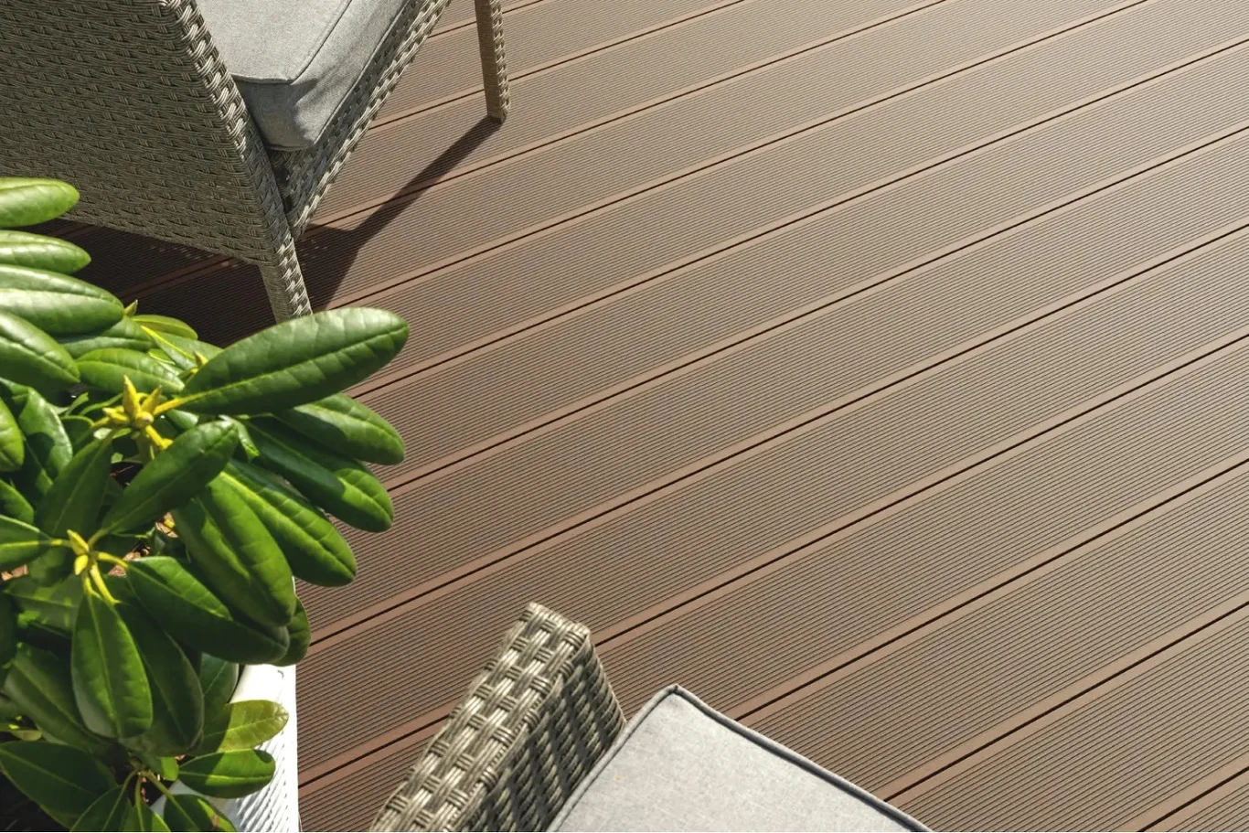 brązowe deski tarasowe o ryflowanej powierzchni, dwa fotele z technorattanu z szarymi poduszkami, w lewym dolnym rogu doniczka z zieloną rośliną o błyszczących liściach