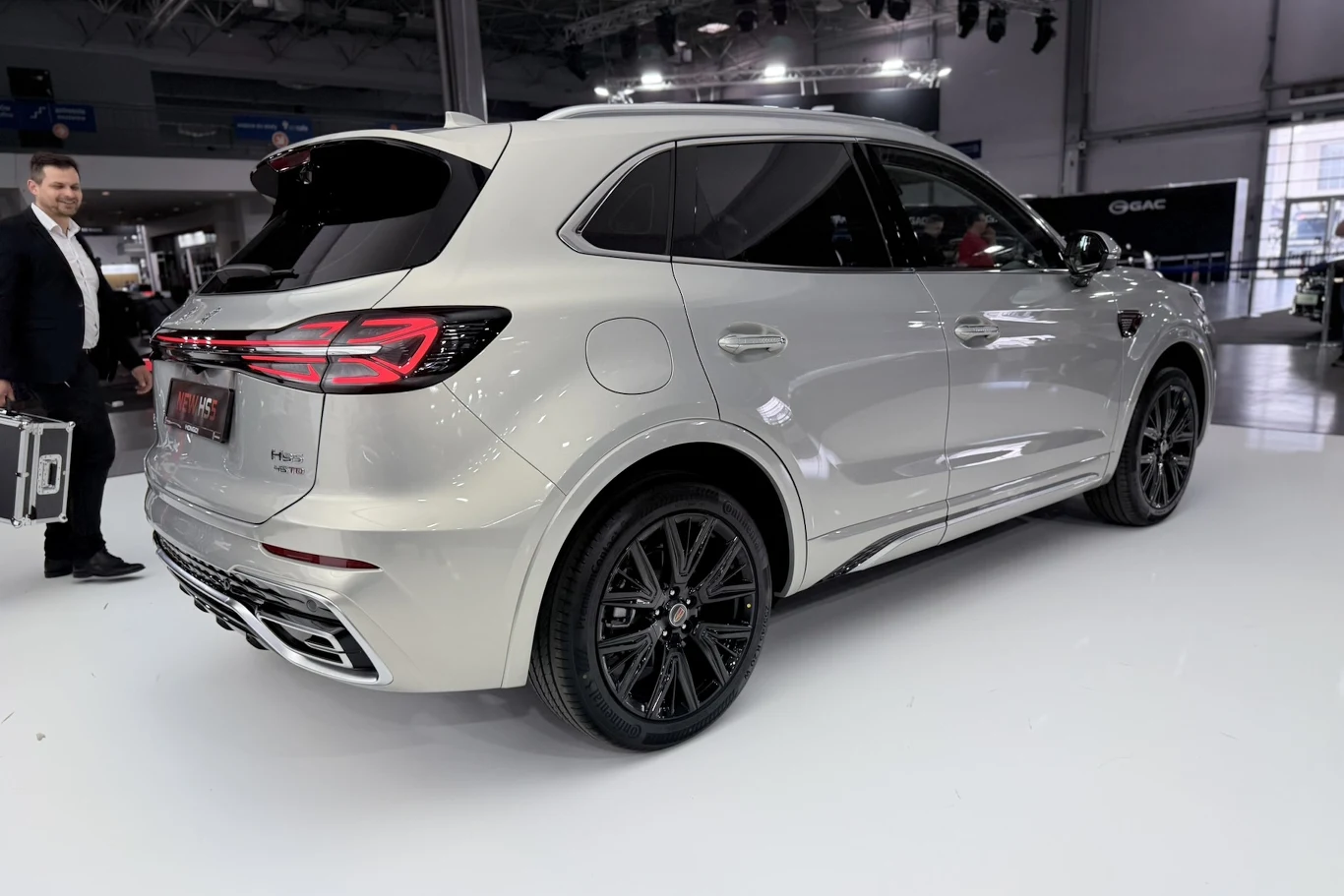 Hongqi HS5 srebrny SUV o nowoczesnym designie, przyciemnianych szybach i czarnych felgach stoi na jasnej hali wystawowej; w tle mężczyzna w garniturze trzymający walizkę